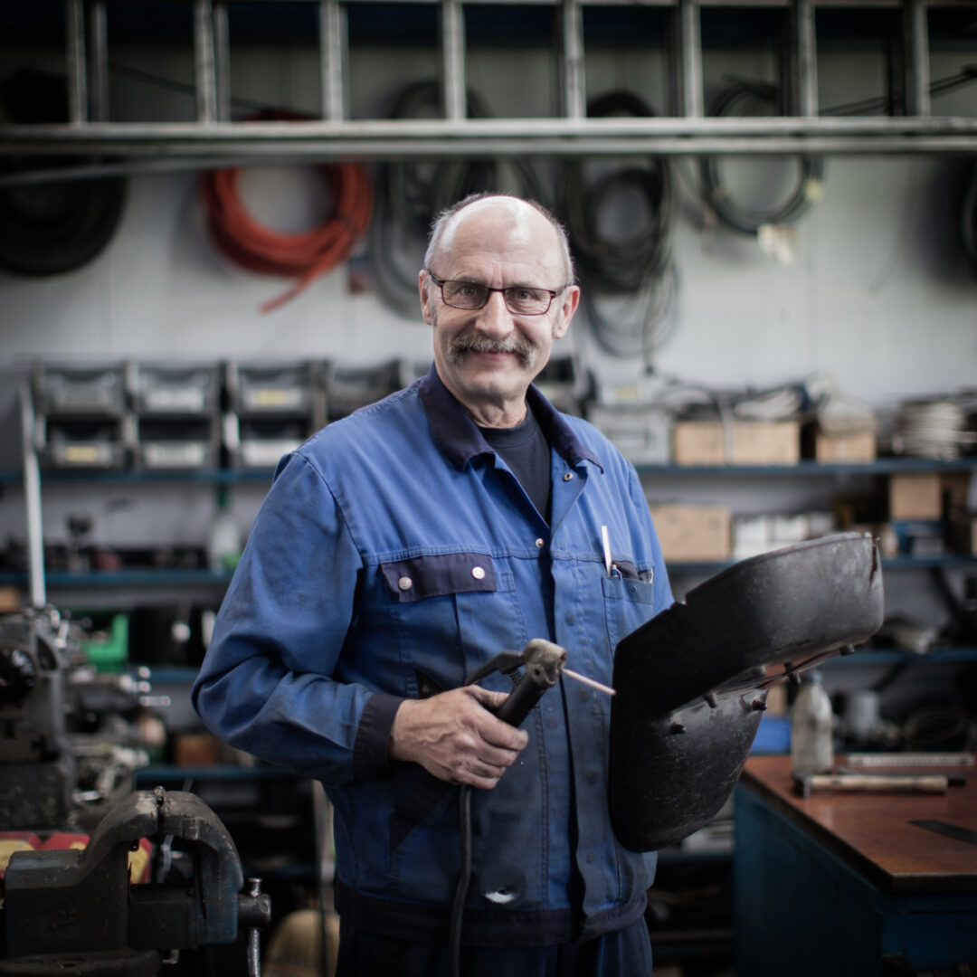 Photo portrait d'un employé qui en habits bleu de travail qui tient dans sa main droite un outil et dans sa main gauche un casque.