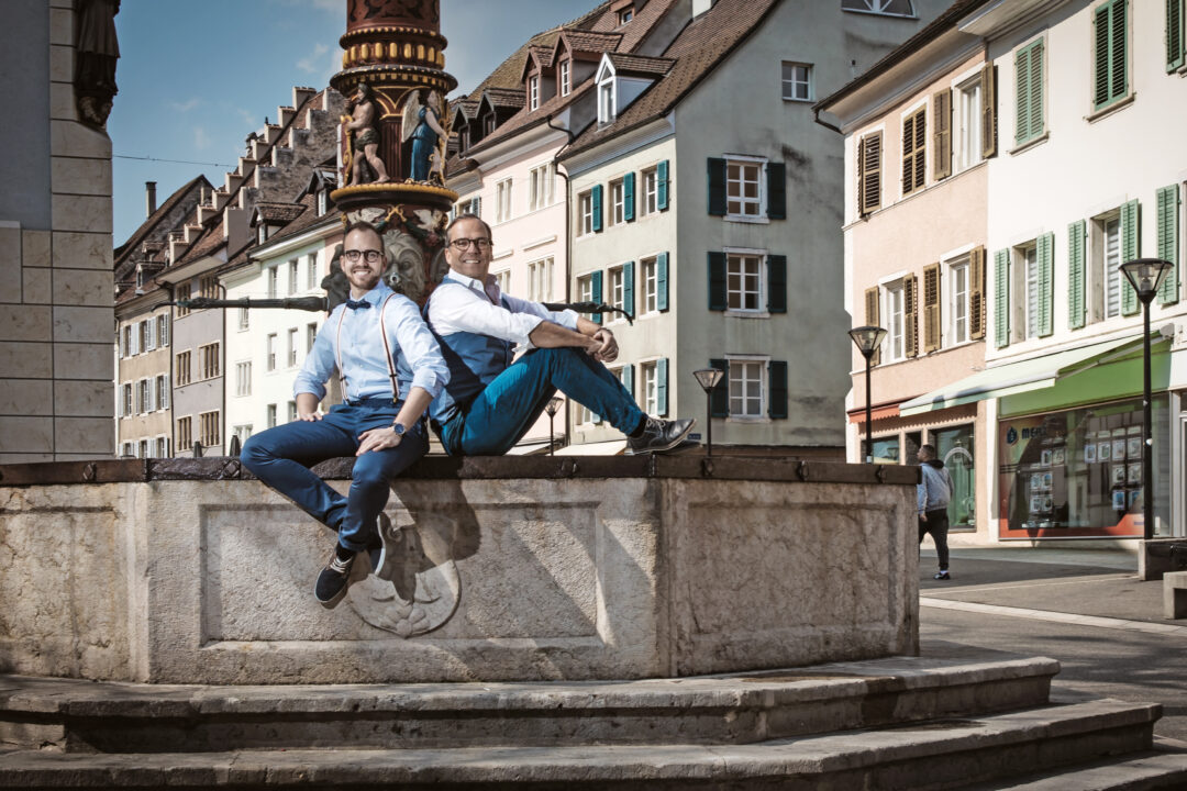 Deux homme portant des lunettes, dos à dos, assis sur le bord d'une fontaine.