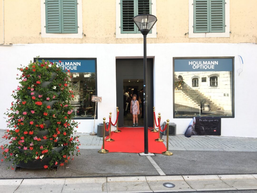 Magasin Houlmann optique vu de dehors, avec un tapis rouge devant l'entrée et un sapin à gauche de l'entrée.